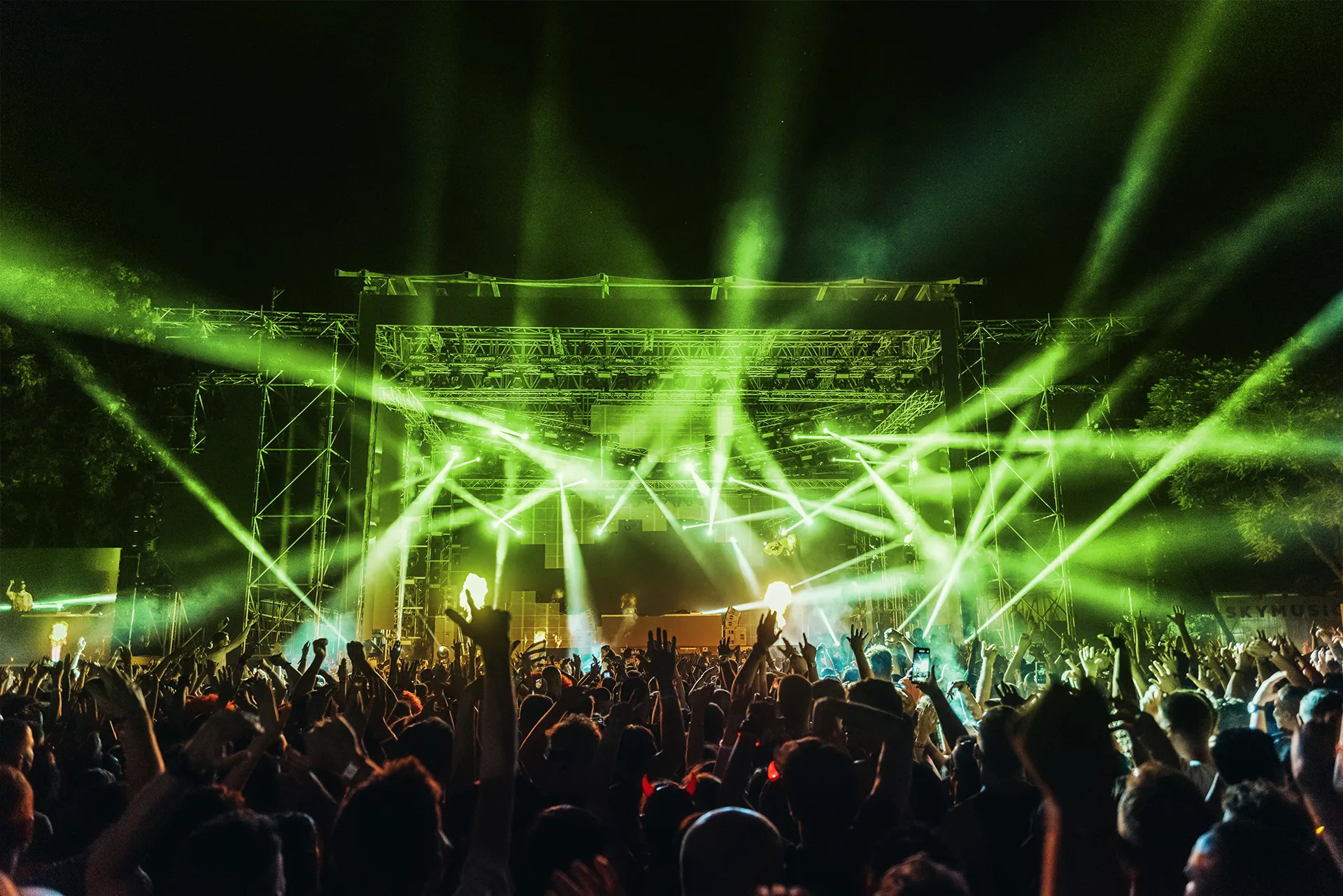 Foule en fête devant une scène illuminée de faisceaux verts lors d’un concert nocturne en plein air.